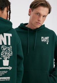 Green hoodie featuring a white printed logo and plant graphic on the back. Includes a front pocket and adjustable drawstring hood.