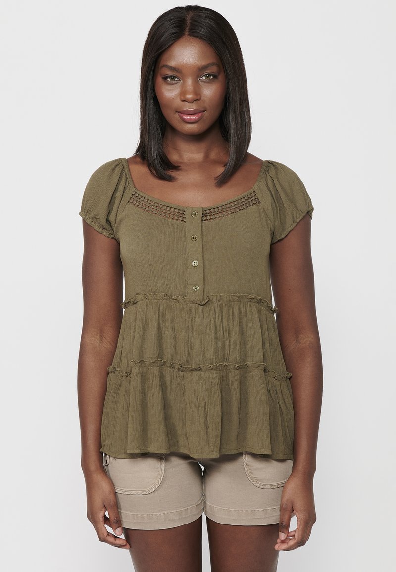 Top peplum verde oliva con maniche corte a sbuffo, chiusura a bottoni e tessuto testurizzato. Orlo a strati con rouches. Indossato con pantaloni corti beige.