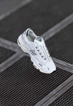 Baskets New Balance 1000 blanches et grises flottant au-dessus d'une surface métallique sombre à motif en grille.