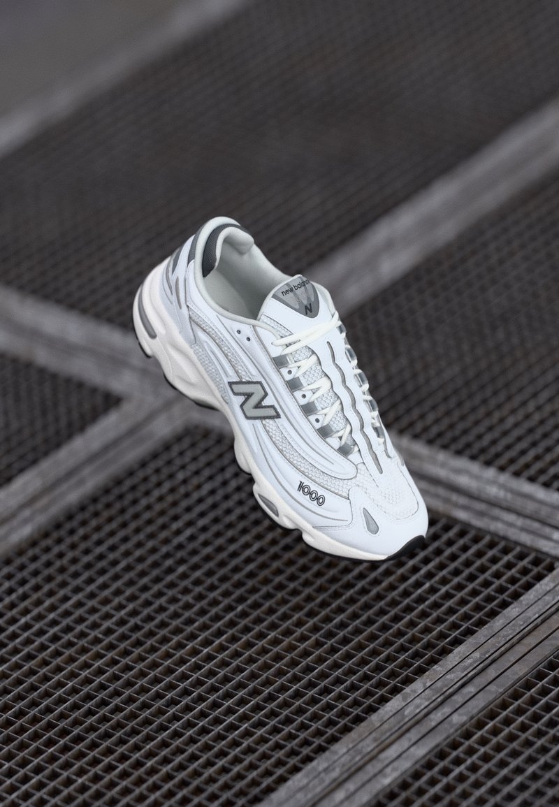 Baskets New Balance 1000 blanches et grises flottant au-dessus d'une surface métallique sombre à motif en grille.