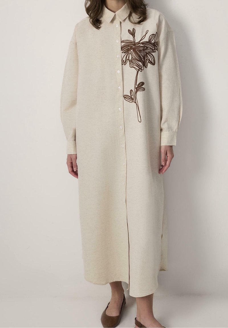 Longue robe beige boutonnée avec col, ornée d'une grande broderie florale marron sur la poitrine droite, portée avec des chaussures plates marron.