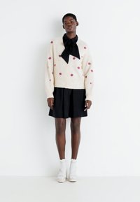 Gilet crème avec des motifs d'étoiles rouges, associé à une robe noire. Le mannequin porte des chaussures à plateforme blanches et des chaussettes montantes, se tenant devant un fond blanc.