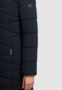 Abrigo tipo plumón azul marino con patrón acolchado, puños elásticos, bolsillo lateral con cremallera y parche de logo en la manga. Tejido suave y ligero.