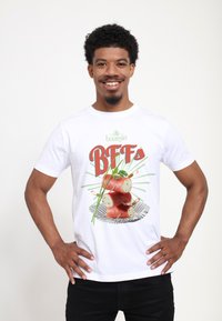 Bílý bavlněný tričko s grafikou stočeného masa a bylinek, na kterém je v zeleném a červeném stylizovaném písmu vytištěno "boursin" a "BFFs".
