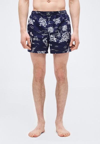 Άνδρας που φοράει σκούρο μπλε μαγιό με λευκό τροπικό και ναυτικό σχέδιο, όρθιος ξυπόλητος μπροστά σε απλό λευκό φόντο.