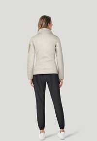 Lys grå, tætsiddende jakke med høj krave og beige skulderlap; parret med sorte joggers og hvide sneakers. Glat tekstur.