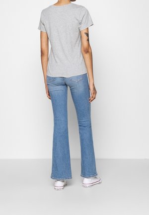 Person steht mit dem Rücken zur Kamera, trägt ein hellgraues kurzärmeliges Hemd, hellblaue ausgestellte Jeans, weiße Turnschuhe und zeigt Tätowierungen an den Armen.