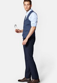 Granatowa kamizelka w marynarskim stylu o teksturowym wykończeniu, sparowana z jasnoniebieską koszulą i ciemnymi spodniami. Brązowe buty wizytowe dopełniają całość.