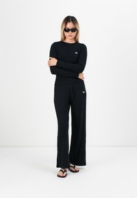 Black ribbed long-sleeve top and wide-leg pants, with a small diamond logo on the top. Model wearing sunglasses and flip-flops.