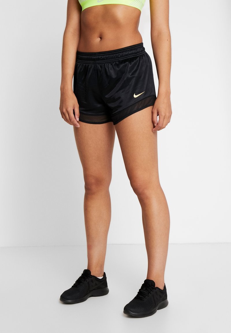 Vrouwelijke torso en benen met zwarte Nike sportshorts, neongele sportbeha en zwarte hardloopschoenen op een witte studiobackdrop.