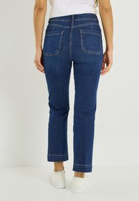 Vaqueros de mezclilla azul oscuro con corte recto, dos bolsillos traseros y un dobladillo limpio. Combinados con zapatillas blancas para un look casual.