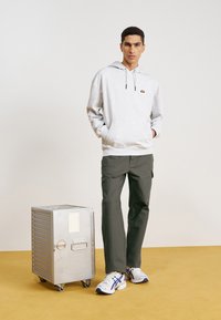 Sudadera gris con capucha y bolsillo, pantalones de cargo verde oliva, y zapatillas blancas con acentos morados, al lado de un contenedor de almacenamiento metálico plateado.