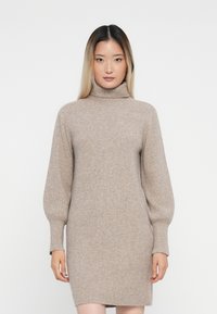 Abito a collo alto in maglia beige con maniche lunghe e gonfiate e texture a coste. L'abito ha una silhouette aderente e arriva sopra il ginocchio.