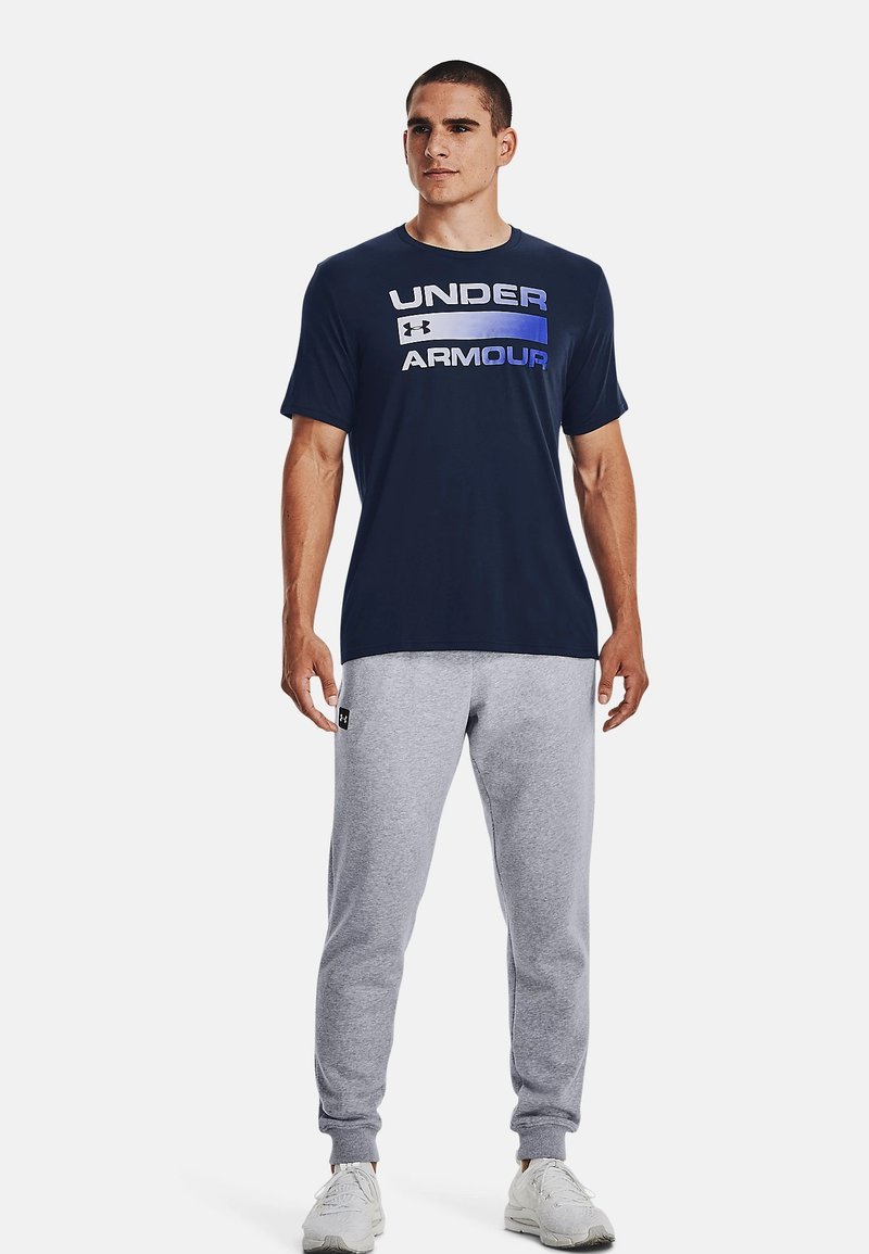 Marinfärgad t-shirt med "UNDER ARMOUR" i vit och blå text, tillsammans med ljusgrå träningsbyxor. T-shirten har rund hals och korta ärmar.