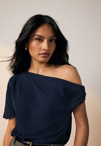 Navy off-the-shoulder top with rolled sleeves, soft texture, and relaxed fit. Worn with a black belt, paired with a neutral background.