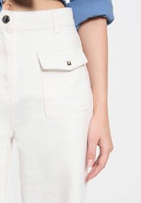 Weiße Denim-Hosen mit einer seitlichen Tasche und goldenen Akzenten sowie einem Knopfverschluss. Die Textur ist glatt mit einer feinen Maserung.