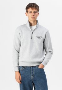 Lysegrå lynlås sweatshirt med høj krave, ribbede mansjetter og kant; har sort logo tekst på venstre bryst; bæres over blå jeans.