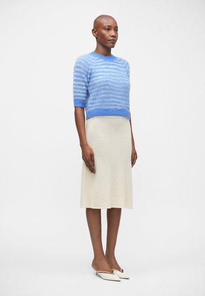 Sweater de manga curta às riscas azuis em tecido suave, combinado com uma saia creme até ao joelho. Sapatilhas brancas de bico fino completam o conjunto.