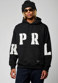 Hoodie noir en coton, décoré de grandes lettres blanches craquelées "PRL" sur le devant et les manches. Comprend une poche avant et une capuche.