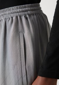 Grå bukser med elastisk midje og diagonalt stikningsmønster, der har en sidelomme og en lille metallisk detalje på midjebåndet.