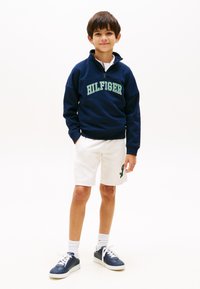 Μπλε σκούρο φούτερ με λευκά και πράσινα γράμματα "HILFIGER", λευκό σορτς με πράσινο λογότυπο και σκούρα μπλε αθλητικά παπούτσια με λευκά κορδόνια.
