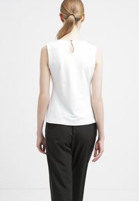 Bílé tílko bez rukávů s průstřihem na zádech, kombinované s černými kalhotami. Hladká látka, jednoduchý design, přiléhavý střih.