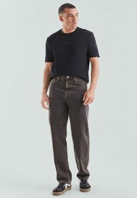 Mann trägt ein schwarzes T-Shirt, ausgewaschene schwarze Jeans und schwarze Sneakers mit weißen Sole und steht vor einem hellgrauen Hintergrund.