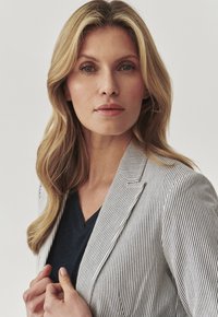 Gestreifter Blazer aus hellgrauem und weißem Stoff, strukturiertes Design, scharfe Revers, getragen über einem dunklen V-Ausschnitt-Top, weiche Textur, natürliche Haare.