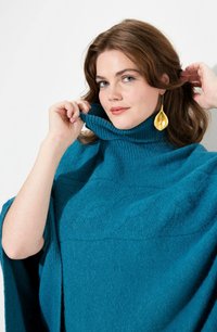 Poncho lavorato a maglia color teal con colletto alto a coste e motivo a treccia. Presentato con orecchini a goccia dorati. Evidente morbidezza della trama.