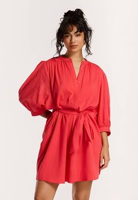 Rode jurk met een losse pasvorm, ballonmouwen en een ceintuur met strik in de taille. Heeft een knoopsluiting met knopen en een zachte, gestructureerde stof.