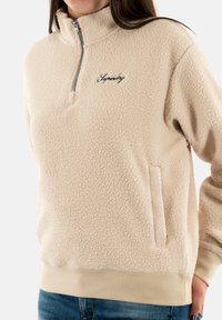 Sudadera de felpa beige con media cremallera, con una superficie texturizada, bolsillos laterales y el logotipo bordado de "Superdry" en el pecho. Puños y dobladillo elásticos.