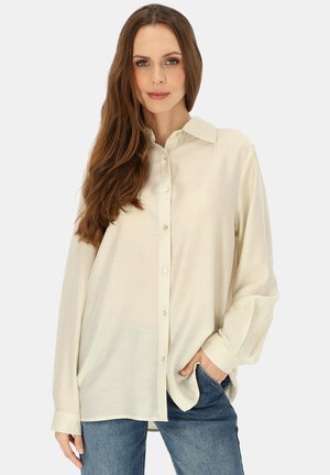 Kobieta z długimi brązowymi włosami, ubrana w kremową koszulę na guziki i niebieskie dżinsy, stoi z jedną ręką w kieszeni na jednolitym tle.