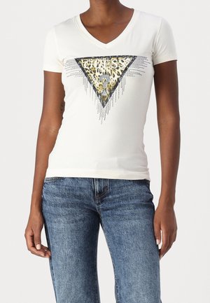 Persoon draagt een wit V-hals T-shirt met een driehoekig logo in luipaardprint met de tekst "GUESS?", gecombineerd met een blauwe spijkerbroek.