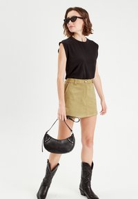 Black sleeveless top, light olive green mini skirt with front pockets, black leather handbag with silver studs, and black cowboy boots.