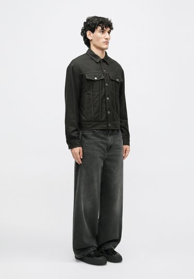 Jachetă din denim neagră cu două buzunare pe piept și închidere cu nasturi, combinată cu blugi negri cu picioare largi și încălțăminte neagră. Textură netedă, design minimalist.