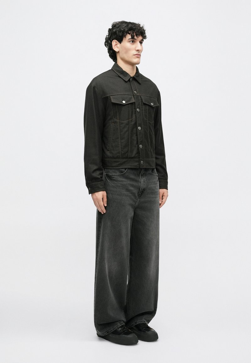 Sort denimjakke med to brystlomme og knaplukning, parret med vide sorte jeans og sorte sko. Glat tekstur, minimalistisk design.