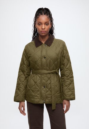 Donna con capelli intrecciati che indossa una giacca trapuntata verde con colletto e cintura marroni, in piedi davanti a uno sfondo bianco uniforme.