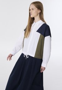 Chemise color-block avec des panneaux blanc, marine et olive, dotée d'un ourlet à cordon de serrage. Des manches longues et un col classique complètent le design.