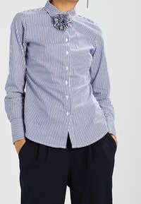 Blue and white striped button-up shirt with long sleeves and a high collar adorned with a fabric flower detail at the neck.
