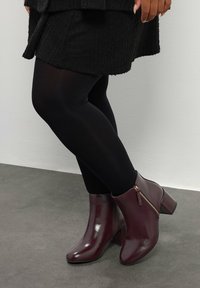 Piernas en pantys negros y botines oscuros burdeos con cremallera lateral, sobre un fondo de suelo gris y pared blanca.