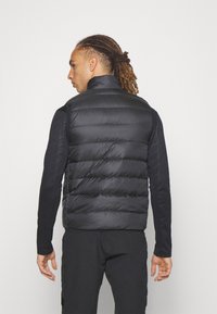 Crni podstavljeni prsluk s podignutim ovratnikom, kvadratnim uzorkom i glatkom teksturom. Nosi se preko crne majice s dugim rukavima i crnih hlača.