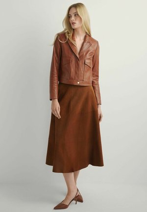 Kobieta z długimi blond włosami ubrana w brązową skórzaną krótką kurtkę, pasującą brązową spódnicę midi oraz brązowe czółenka ze szpiczastym noskiem i paskiem na pięcie, stoi.