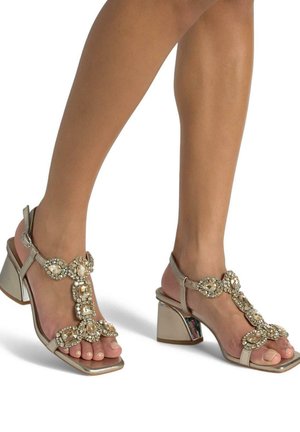 Sandali con tacco metallici beige con design a T-strap, decorati con grandi gioielli trasparenti e punta quadrata. Tacco largo e lucente.