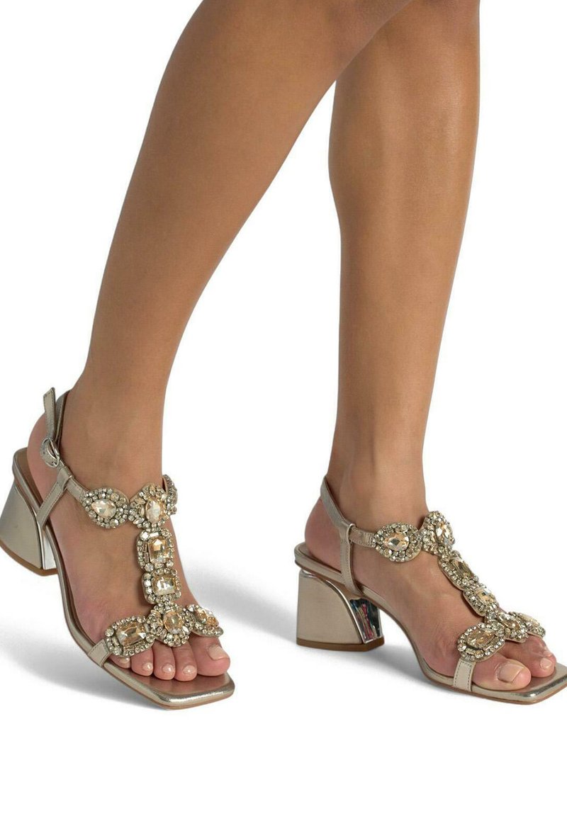 Sandali con tacco metallici beige con design a T-strap, decorati con grandi gioielli trasparenti e punta quadrata. Tacco largo e lucente.