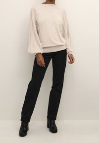 Heller beiger Pullover mit gerippten Ballonärmeln, weichem Material und schmalem Saum, kombiniert mit schwarzen lässigen Jeans und Stiefeletten mit Blockabsatz.