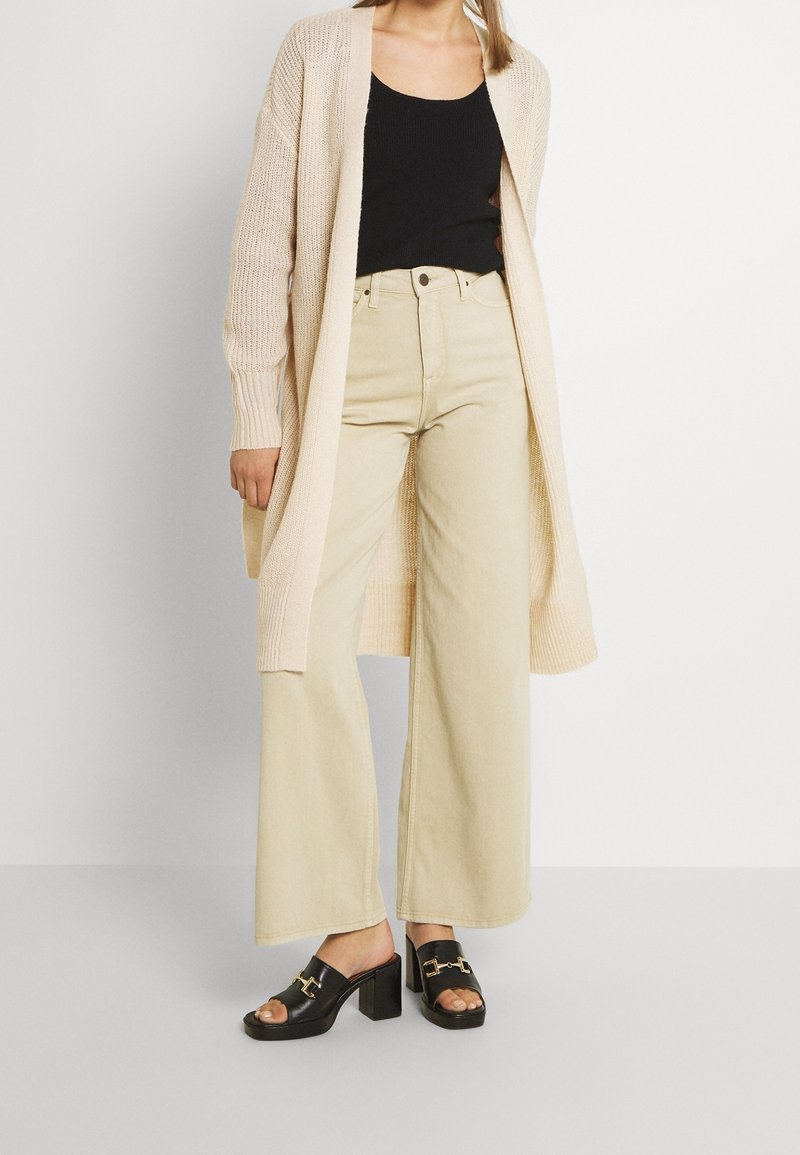 Cardigan long beige en tricot porté sur un débardeur noir, associé à un pantalon beige taille haute à jambes larges et des sandales à talons noires avec des accents dorés.