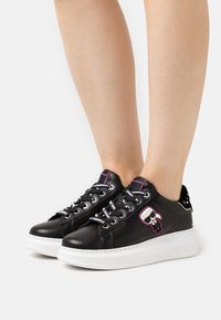 Černé kožené tenisky s bílou podrážkou, které mají grafiku postavy, barevné tkaničky a cvočky na patě.