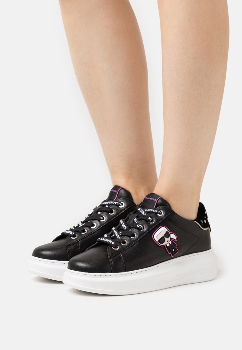 Černé kožené tenisky s bílou podrážkou, které mají grafiku postavy, barevné tkaničky a cvočky na patě.