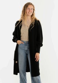Jonge vrouw met lang blond haar, gekleed in een beige top, een zwarte lange cardigan en lichtblauwe jeans, die glimlacht en iets naar rechts kijkt.