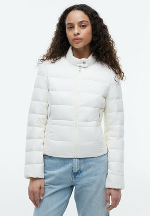 Kvinde med langt krøllet hår iført en hvid quiltet jakke med høj krave og lyseblå jeans, står mod en ensfarvet baggrund.